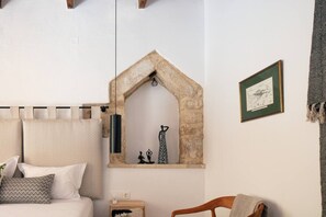 A bedroom with a bed, a nightstand, a wooden chair, a stone arch window, and a framed picture on the wall.