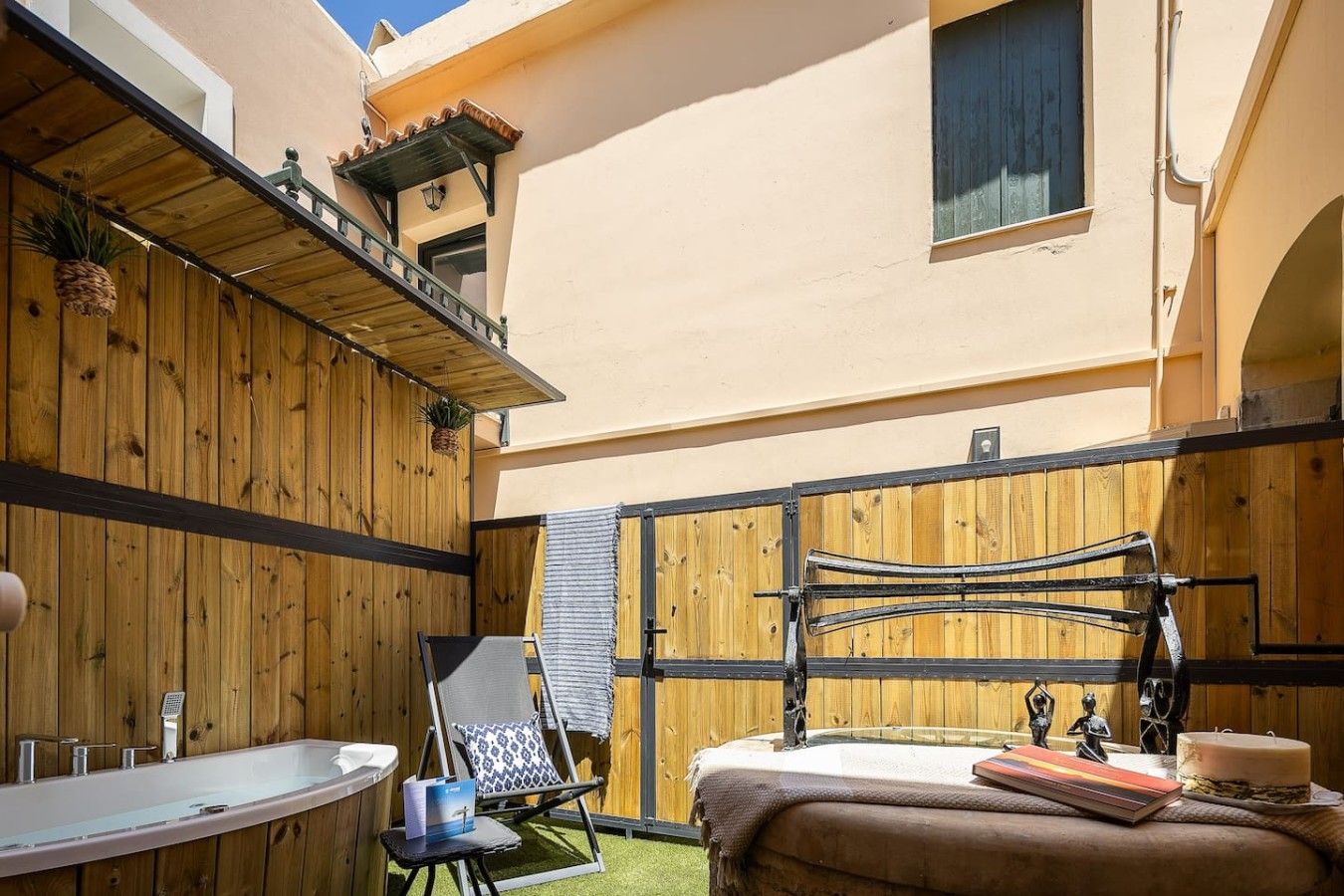 A sunlit outdoor patio with a wooden deck, a lounge chair, a small table with a book and a cake, and a window with a view of the building.