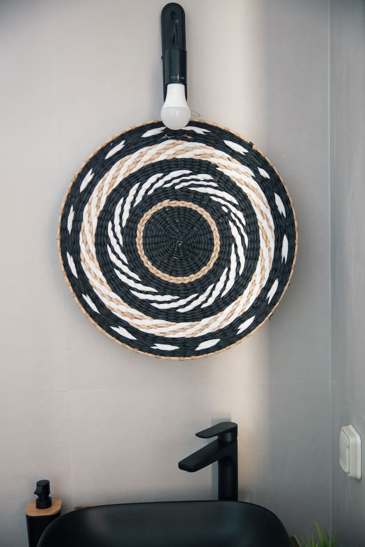 Un tapiz circular tejido con un diseño blanco y negro, un grifo negro y un taburete de madera.