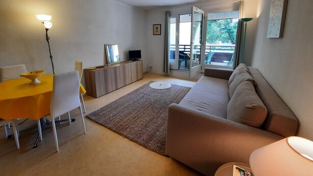 A compact living space with a dining area, a sofa, a TV on a wooden cabinet, and a balcony with a view of greenery. - Beachside Retreat: Cozy 1BR Apt with Balcony, WiFi & Parking (Seignosse)