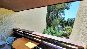 A balcony with a wooden table and two chairs, a white plastic chair, and a view of greenery and parked cars. - Beachside Retreat: Cozy 1BR Apt with Balcony, WiFi & Parking (Seignosse)