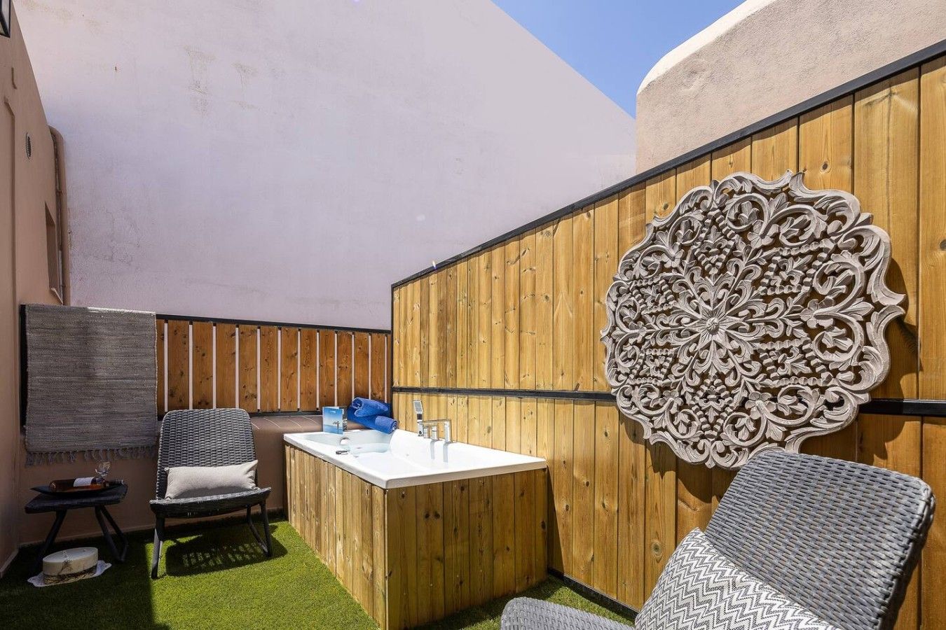 A small outdoor area with a wooden bench, a chair, a table with a sink, and a decorative wall panel.