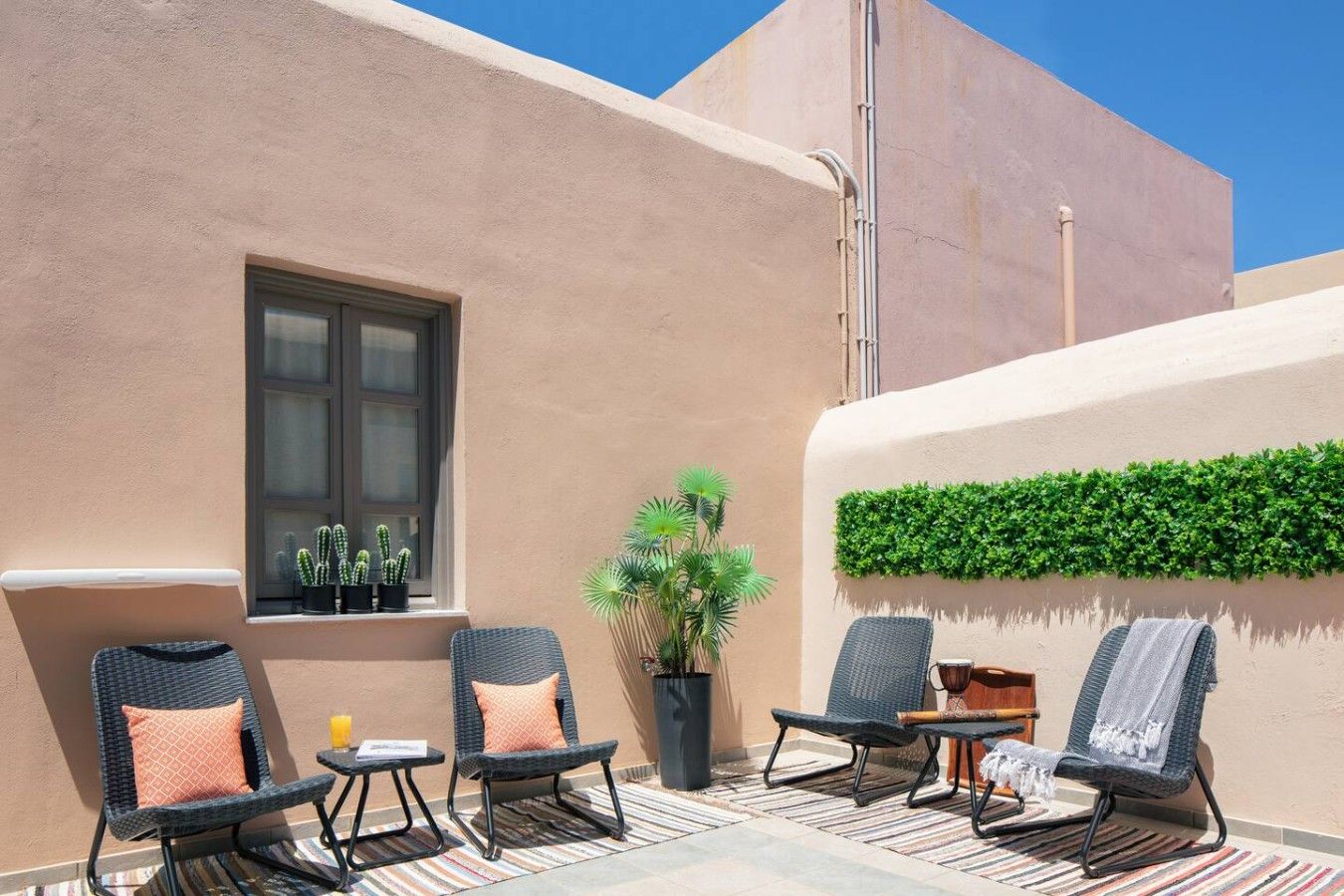 An outdoor seating area with wicker chairs and cushions, a window with potted plants, and a wall-mounted green plant.