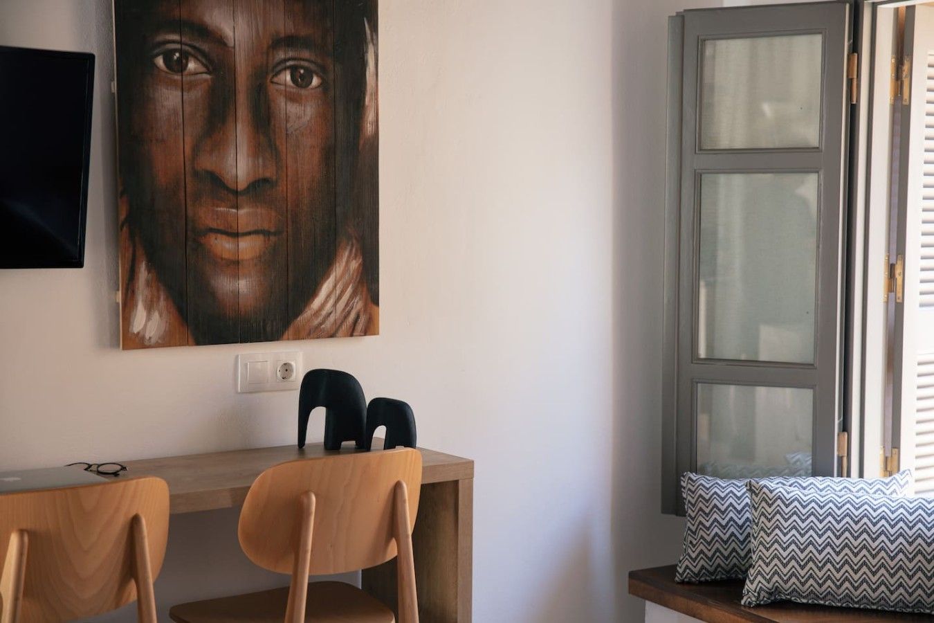 Ein Zimmer mit einem Holztisch, zwei Stühlen, einem an der Wand montierten Fernseher und einem Fenster mit Jalousien.
