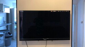 Un téléviseur à écran plat est fixé sur un mur blanc dans une pièce qui comprend une porte, une poubelle et une petite table.
