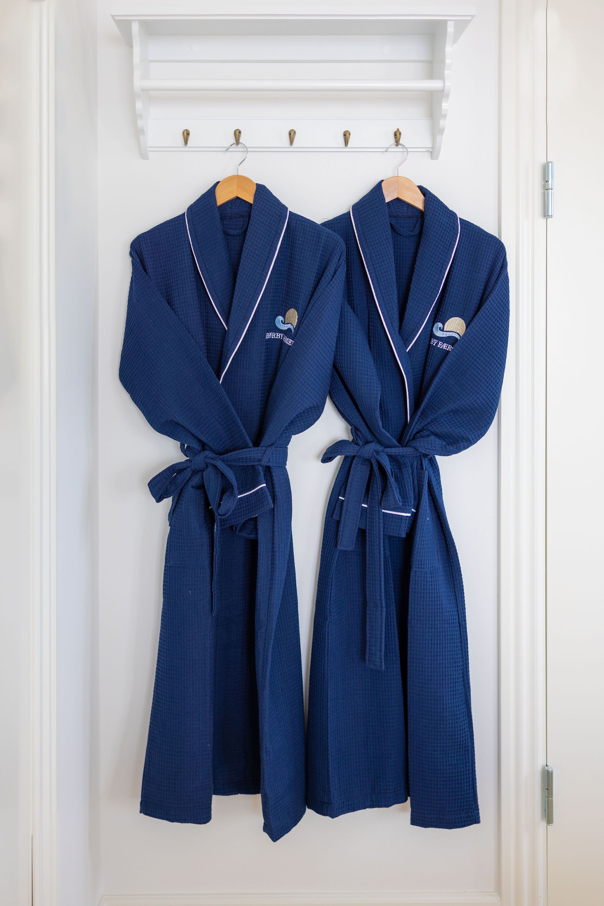 Two blue robes hanging on a white door with a white shelf above them.