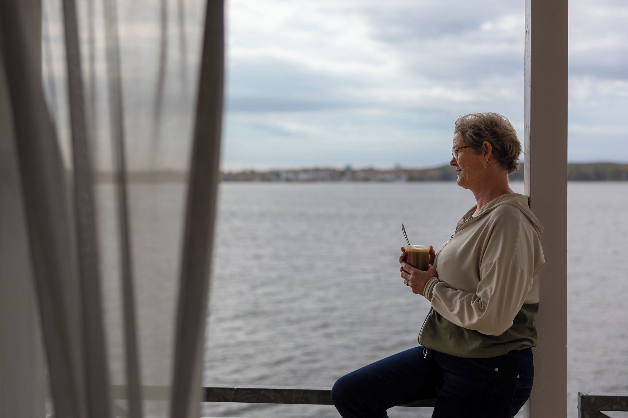 A person sitting by a window overlooking a body of water, holding a cup.