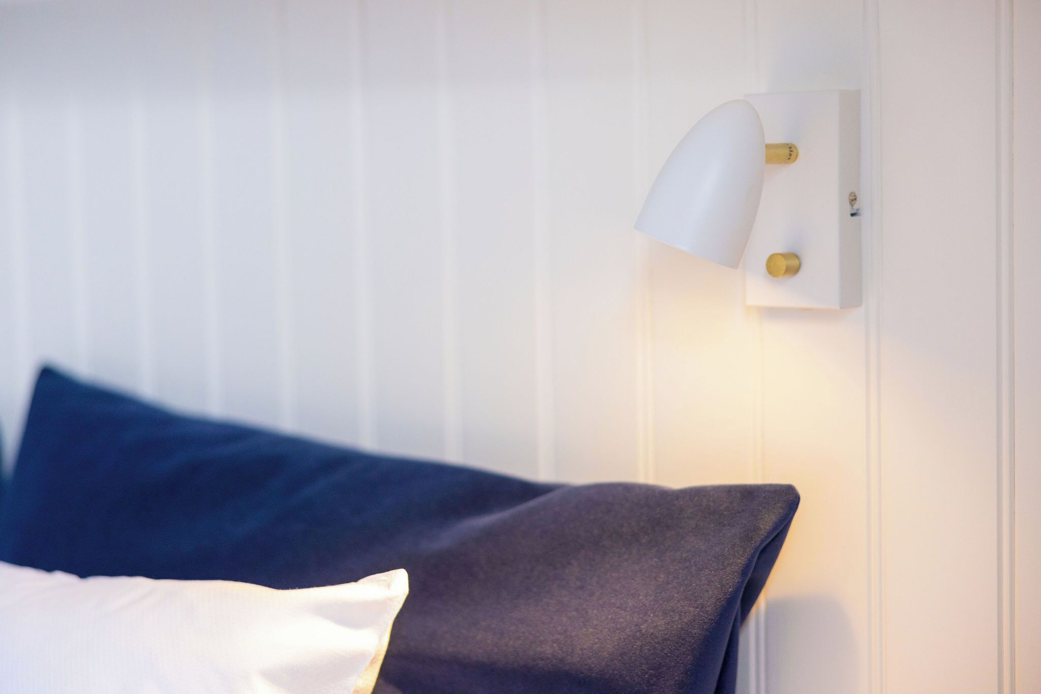Ein Bett mit weißer und dunkelblauer Bettwäsche, eine weiße Wand und eine an der Wand befestigte Lampe.
