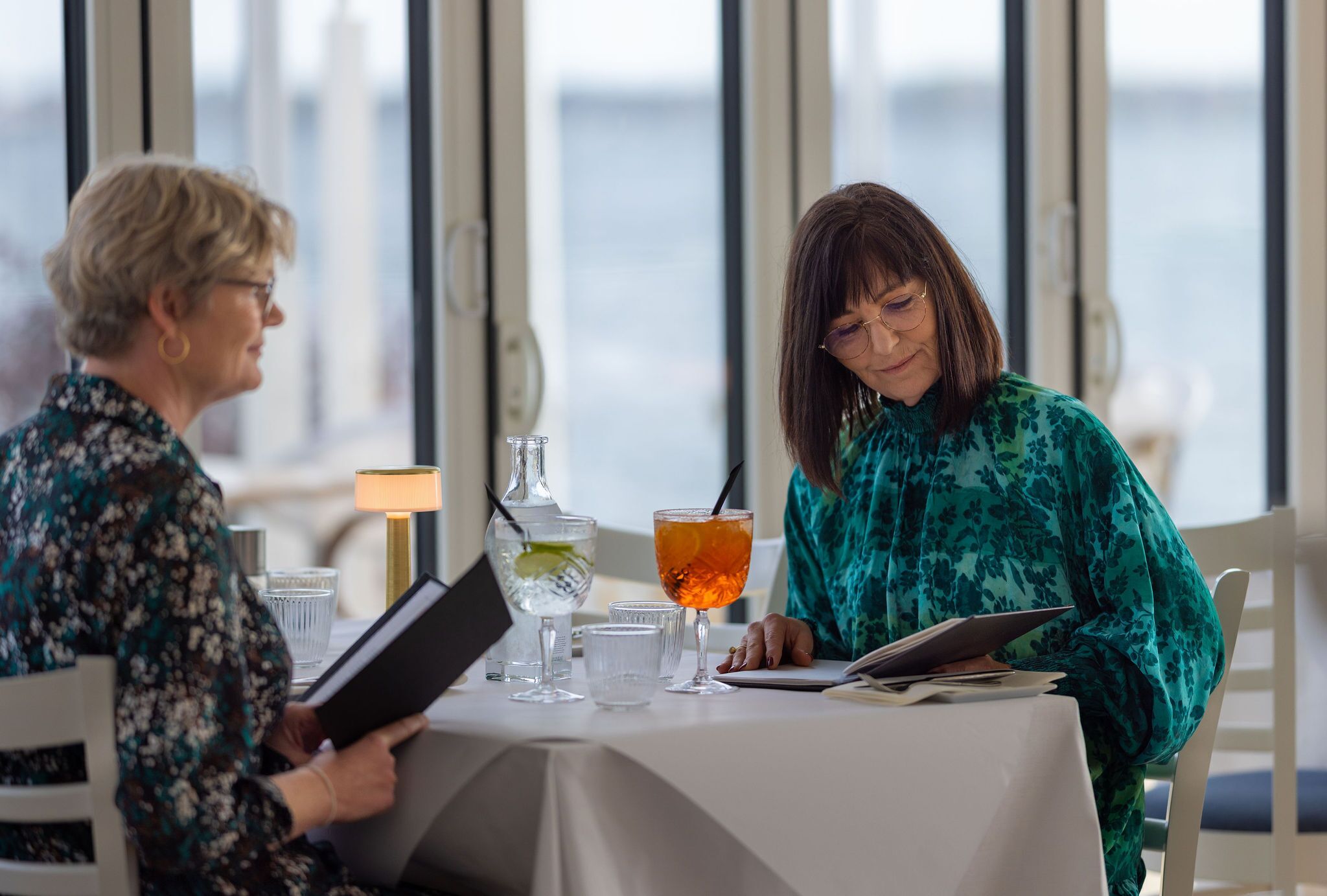 Dos personas sentadas en una mesa con vistas al mar, un vaso de agua y una bebida sobre la mesa.