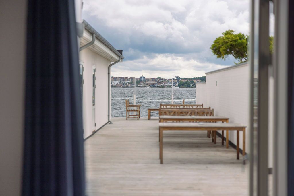 A view from a room with a wooden bench and chairs facing a body of water, with buildings in the background.