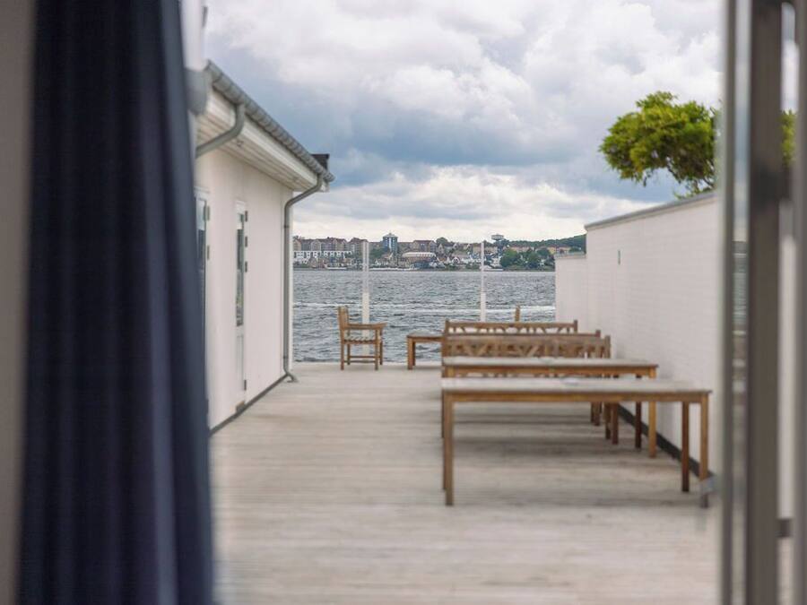 A view from a room with a wooden bench and chairs facing a body of water, with buildings in the background.