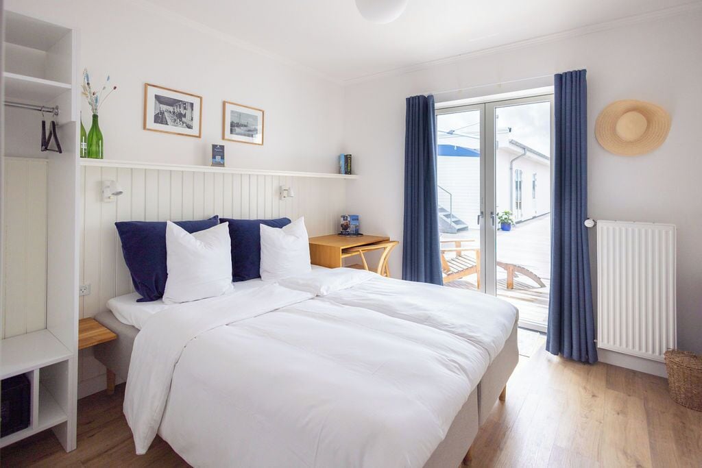 Un dormitorio con cama, una mesita de noche de madera, una pequeña mesa, una ventana con cortinas y un radiador.