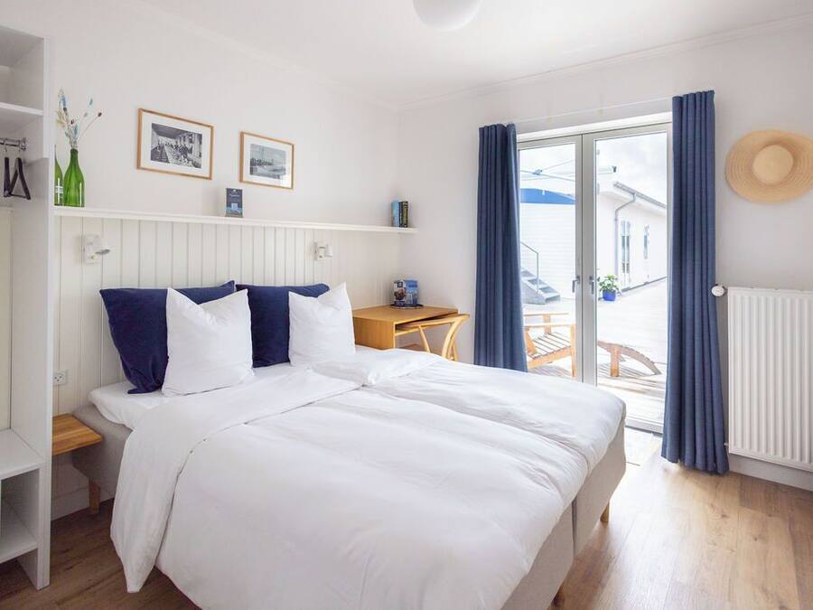 Un dormitorio con cama, una mesita de noche de madera, una pequeña mesa, una ventana con cortinas y un radiador.