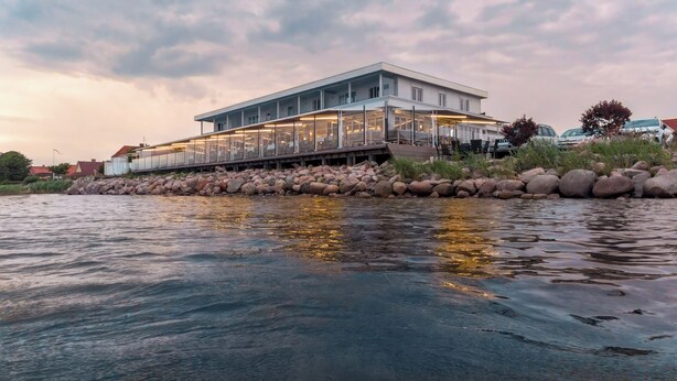 A modern building with a glass facade, a deck, and a waterfront setting. - Hørby Færgekro (Holbaek)