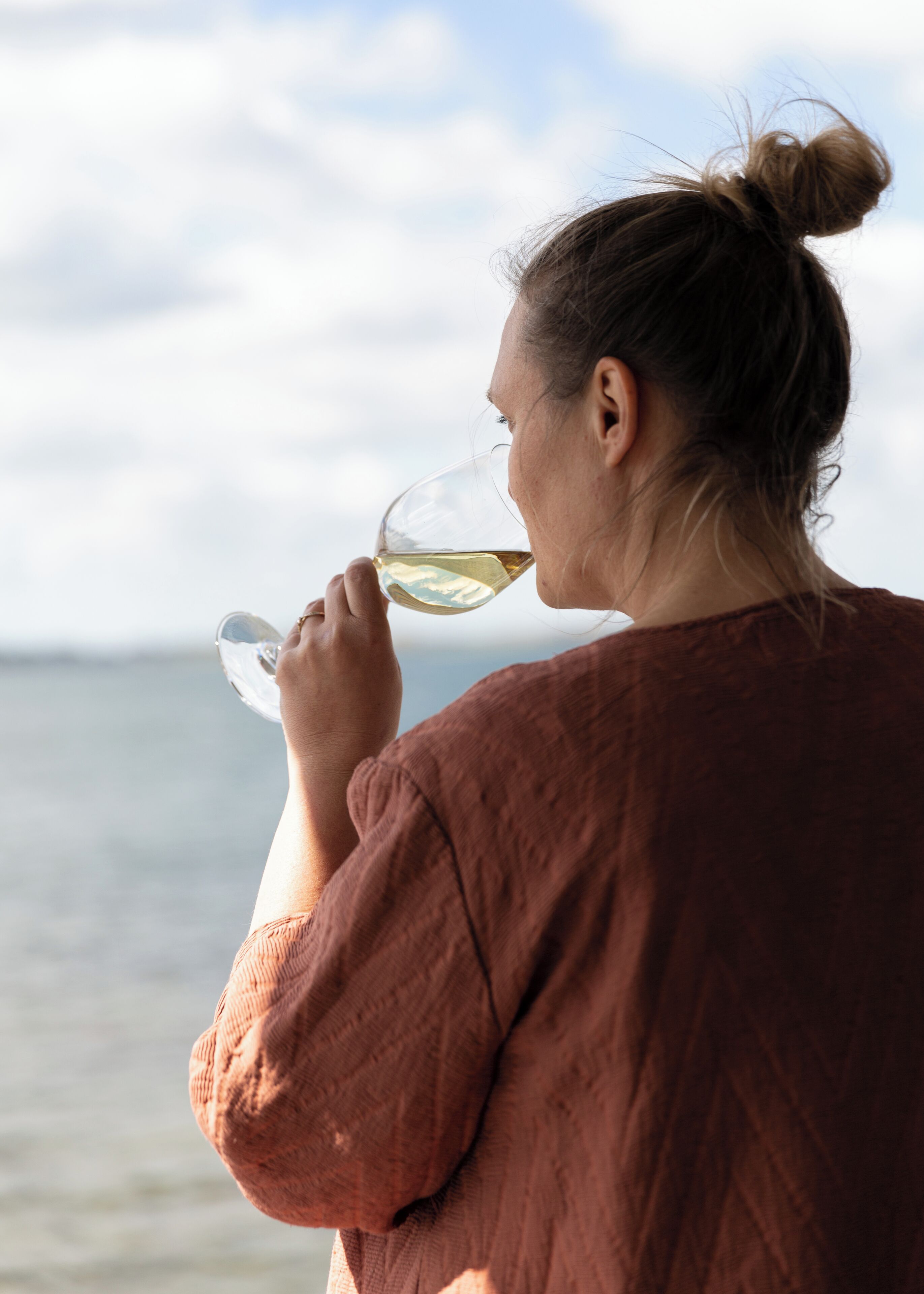 A person holding a glass of white wine, wearing a brown top, with a body of water and a cloudy sky in the background.