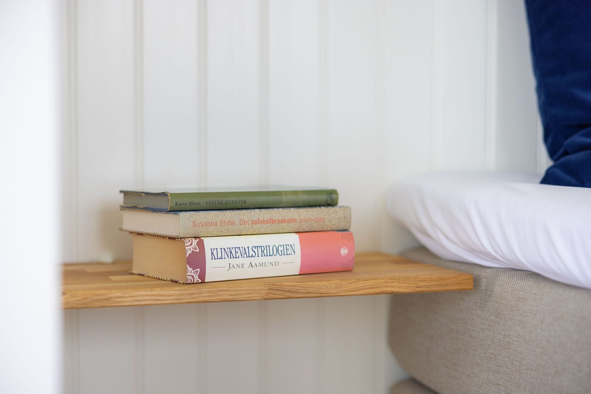 Una pila de libros sobre un estante de madera junto a una cama con un almohadón blanco.