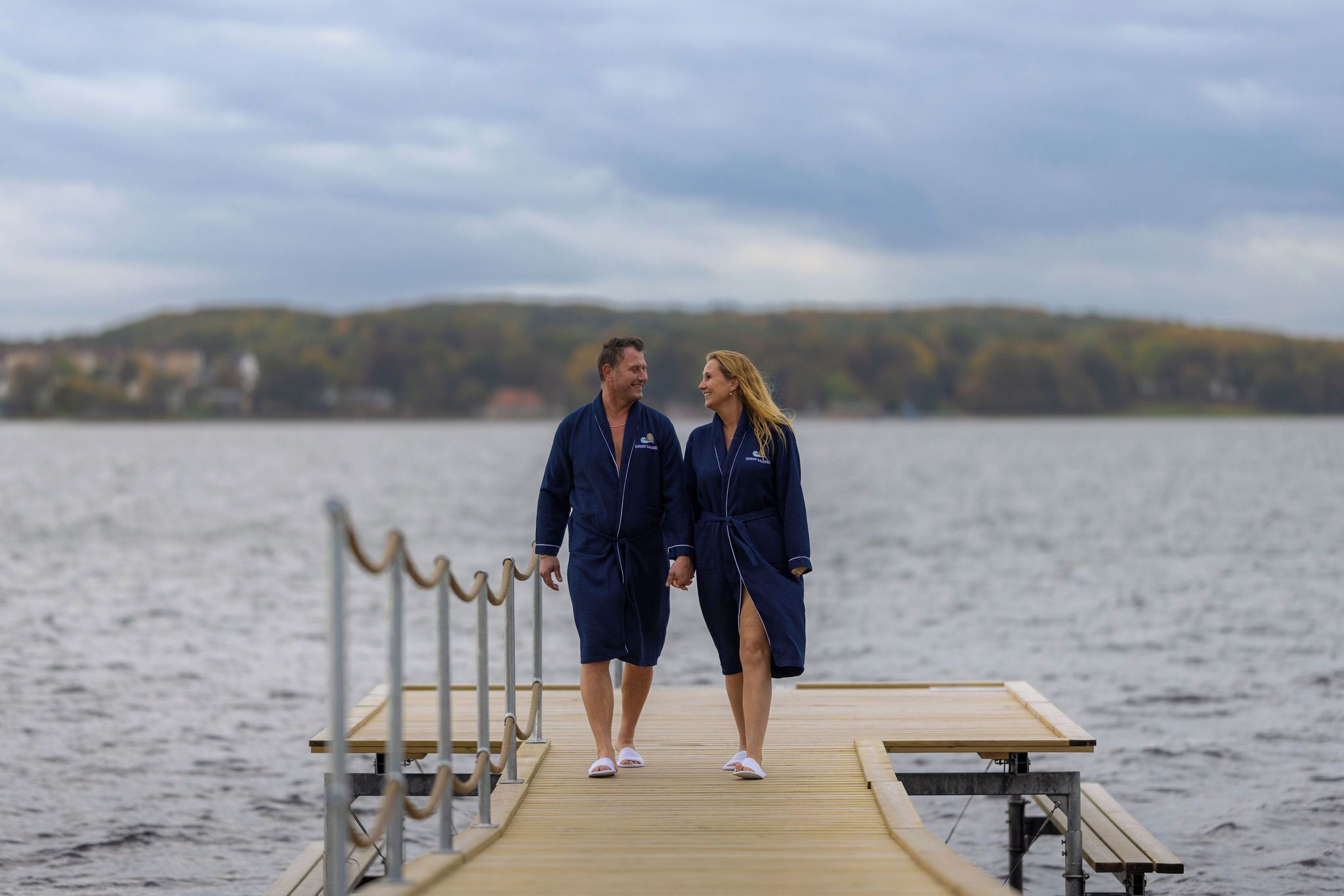 Two people walking on a wooden dock over water with a hilly landscape in the background.