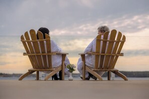 Deux fauteuils Adirondack en bois, placés face à face, avec un vase de fleurs entre eux, et offrant une vue sur une étendue d’eau.