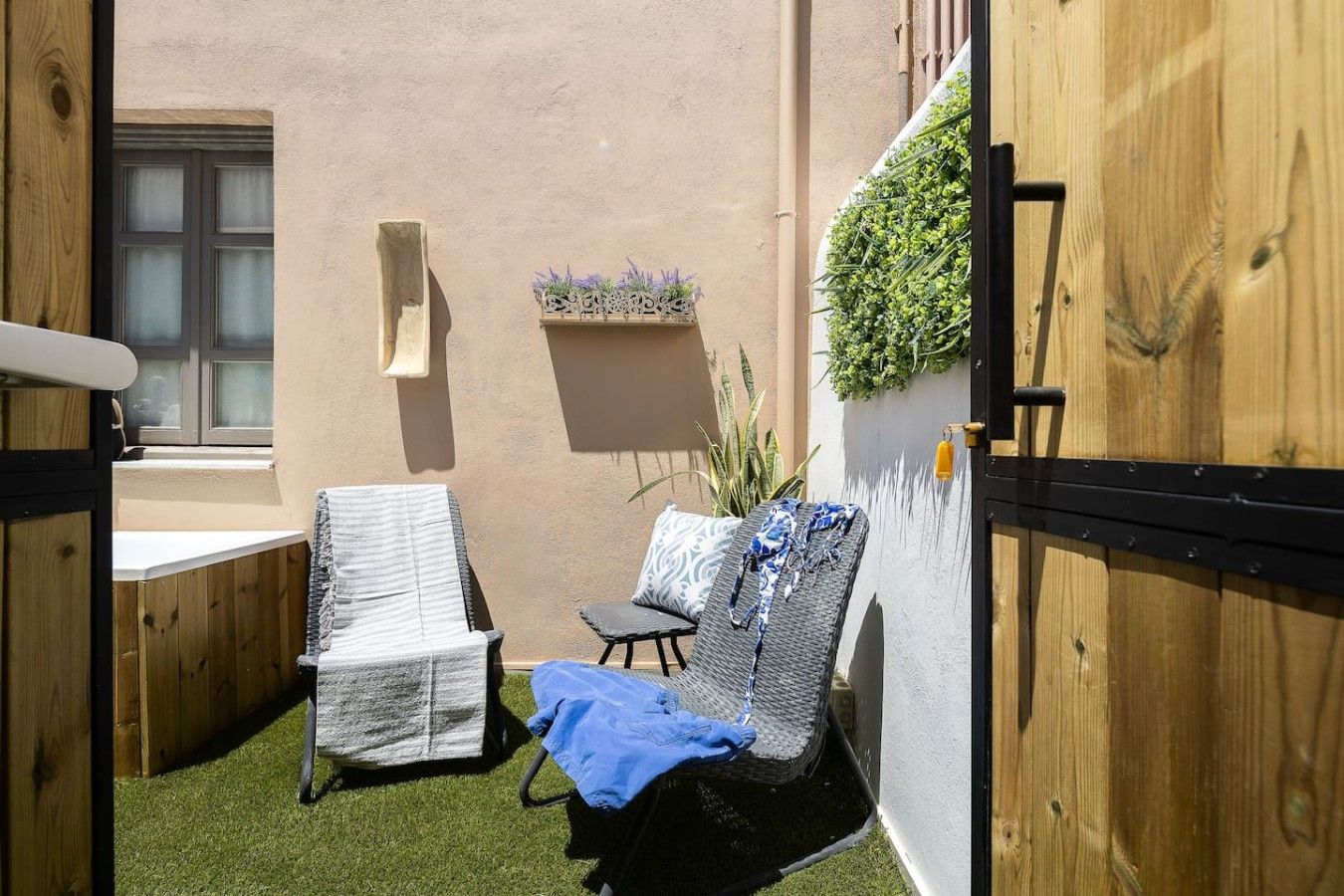 A small outdoor seating area with two lounge chairs, a wooden door, and a wall-mounted planter with greenery.