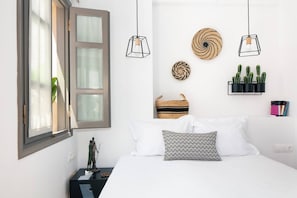 Un dormitorio con una cama, una mesita de noche, un estante en la pared con libros y una planta, y una ventana con cortinas.