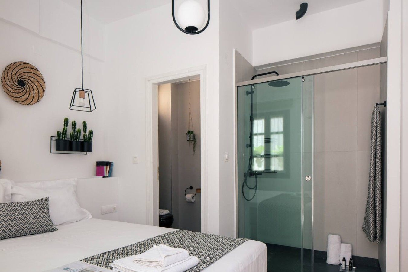Un dormitorio moderno con cama, mesitas de noche, lámpara colgante, zona de ducha con puertas de cristal y un baño pequeño.