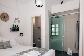 A modern bedroom with a bed, bedside tables, a hanging light fixture, a shower area with glass doors, and a small bathroom. - Gateway Suites - Pathos (Ρέθυμνο)