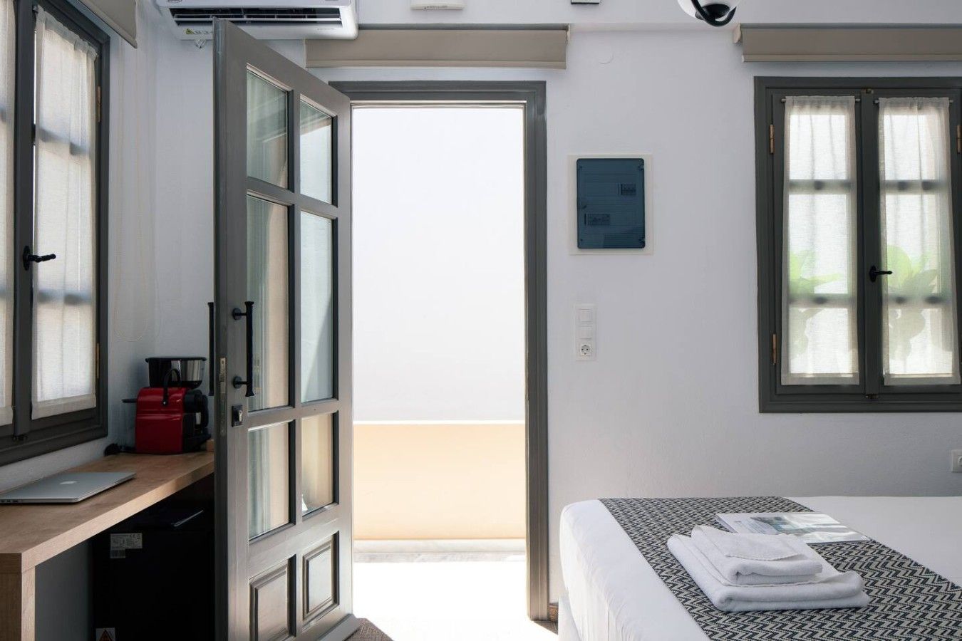 A hotel room with a bed, a desk with a laptop, a red kettle, and a view of the outside.