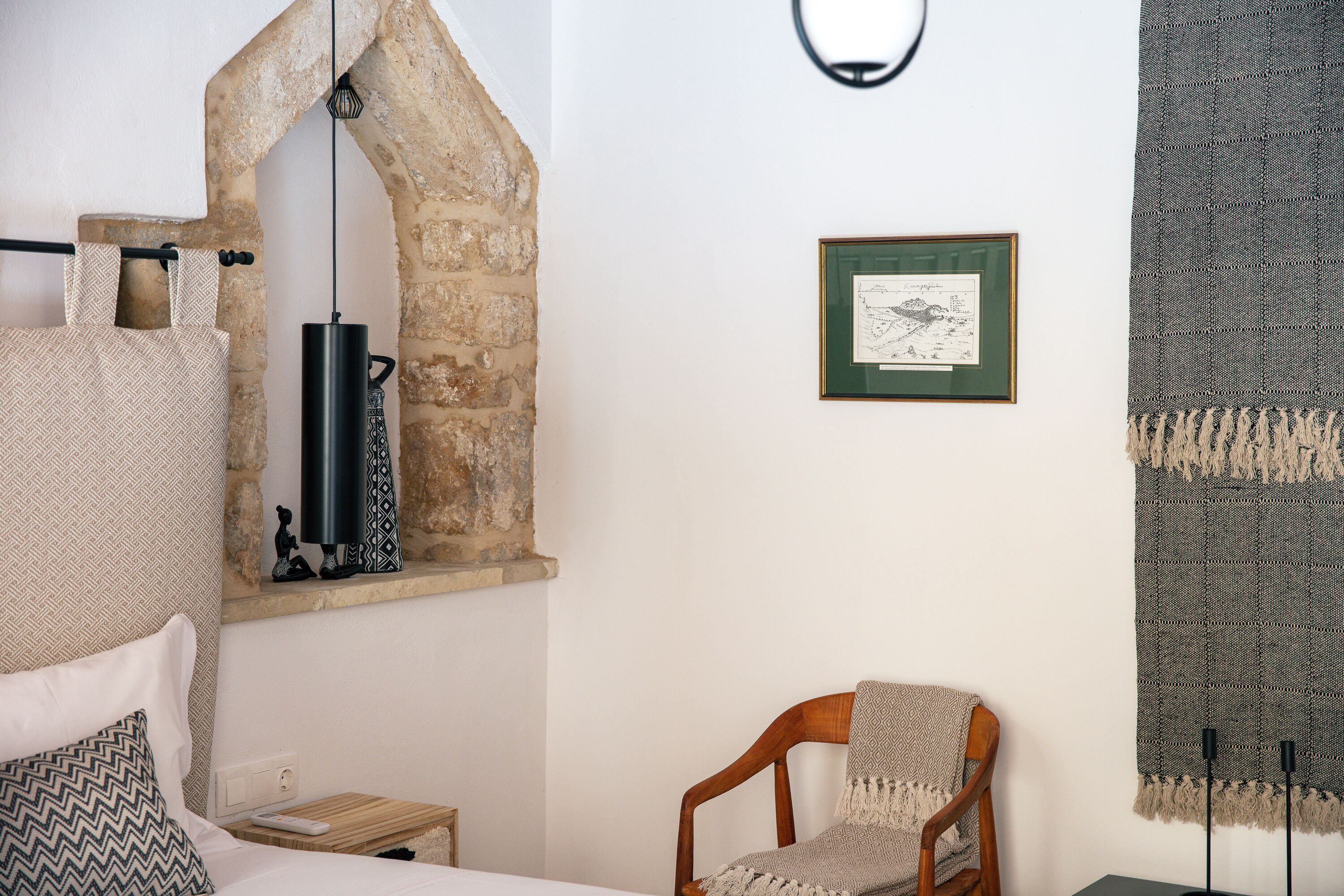 Ein Schlafzimmer mit einem Fenster in Form eines Rundbogens aus Stein, einem Bett mit gemusterten Kissen, einem Holzstuhl, einem kleinen Nachttisch und einem gerahmten Bild an der Wand.