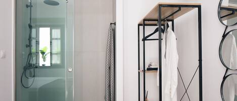 A modern bathroom with a glass shower enclosure, a wooden shelf unit with hanging towels, and a wall-mounted mirror.