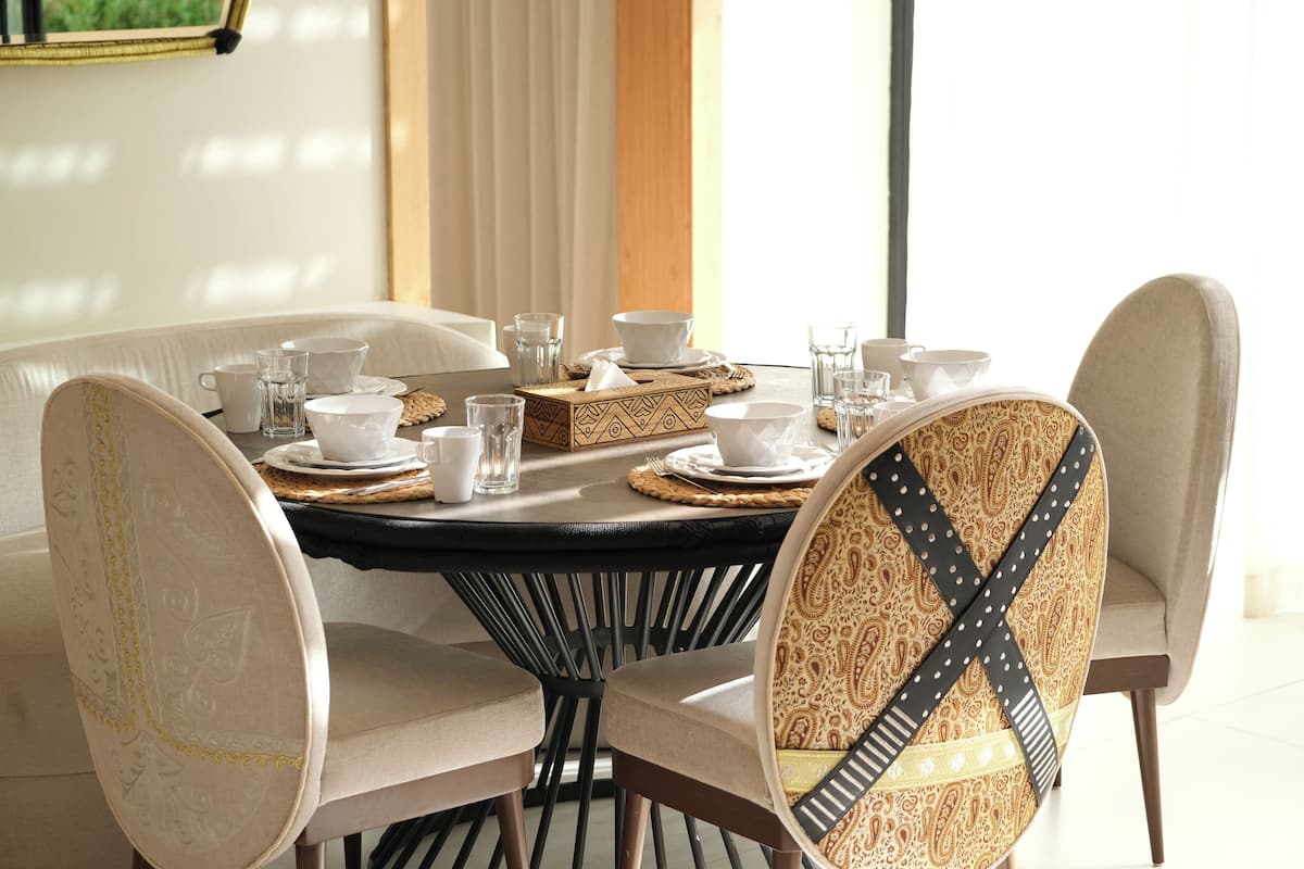 A dining area with a round table set for four, featuring a black tablecloth, white plates, and beige chairs with patterned upholstery.