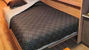 Un lit compact agrémenté d’une couvre-lit à motifs géométriques, d’une tête de lit en bois et d’une petite plante en pot posée sur une étagère au-dessus.