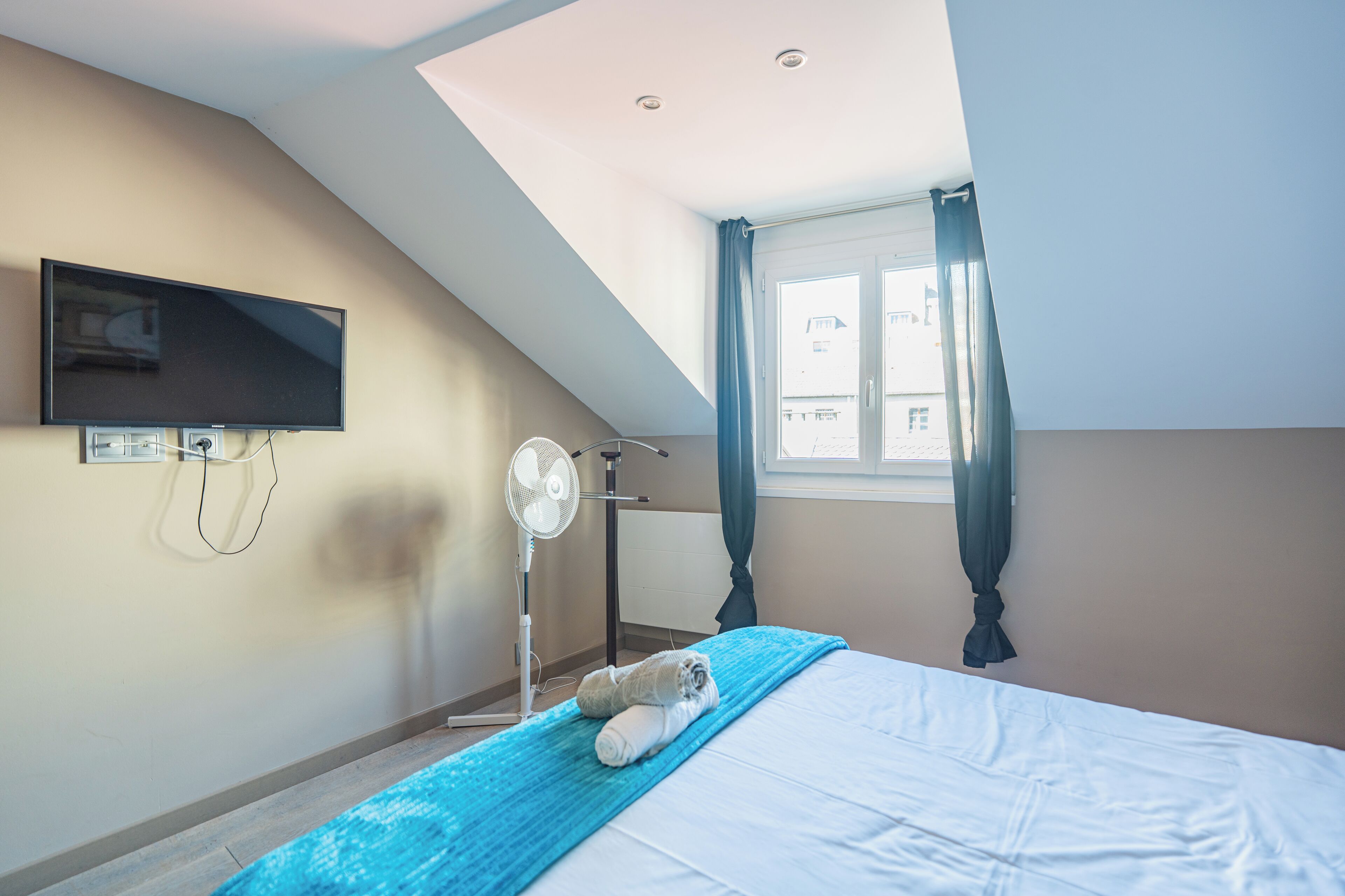 A bedroom with a bed, a TV mounted on the wall, a standing fan, and a window with curtains.