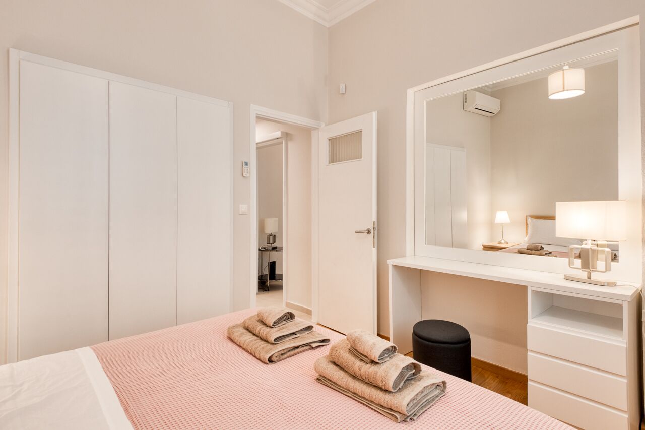 A hotel room with a bed, a desk with a lamp, a mirror, and a small trash bin.