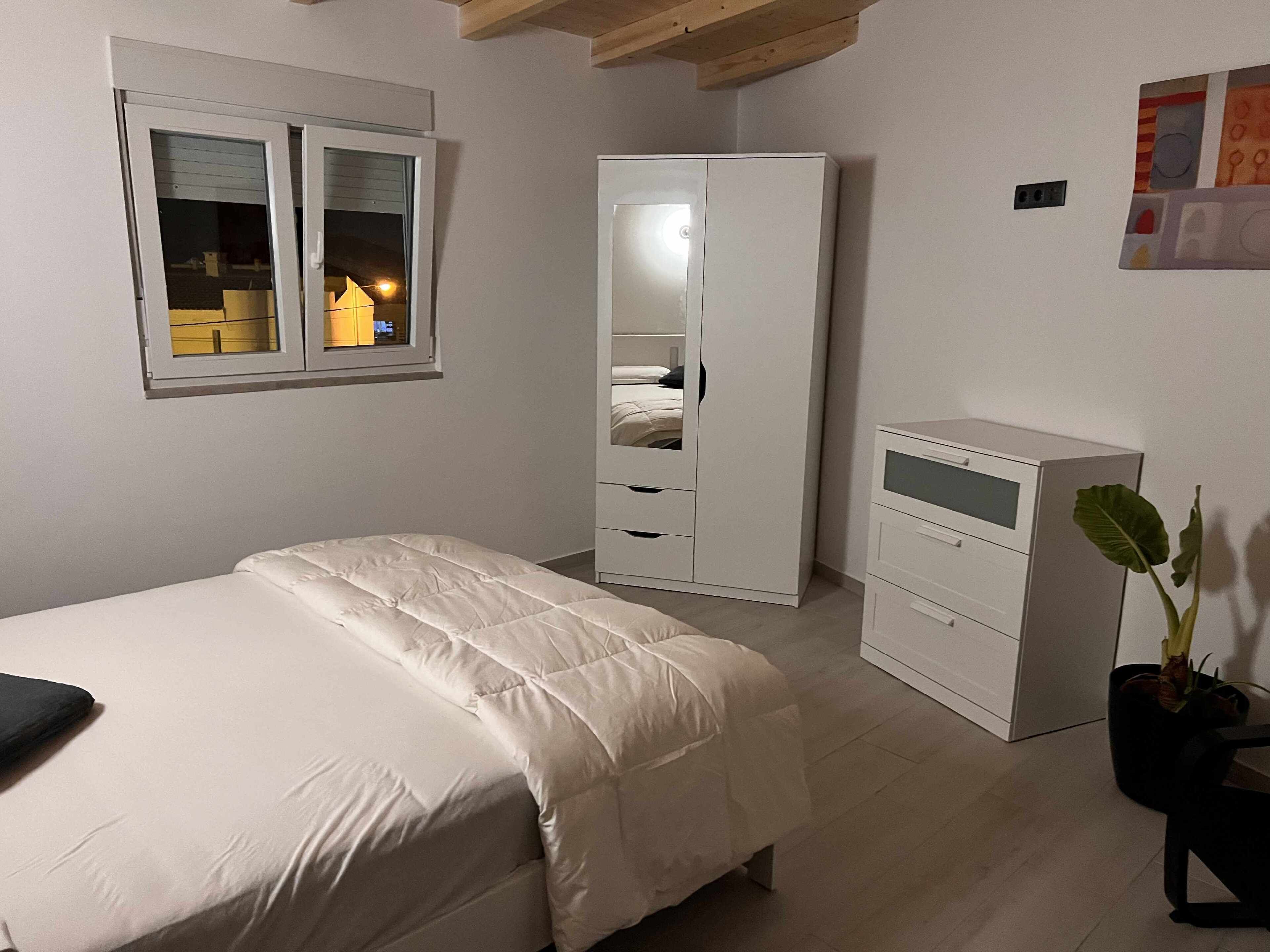 A bedroom with a bed, a wardrobe, a desk, and a potted plant.