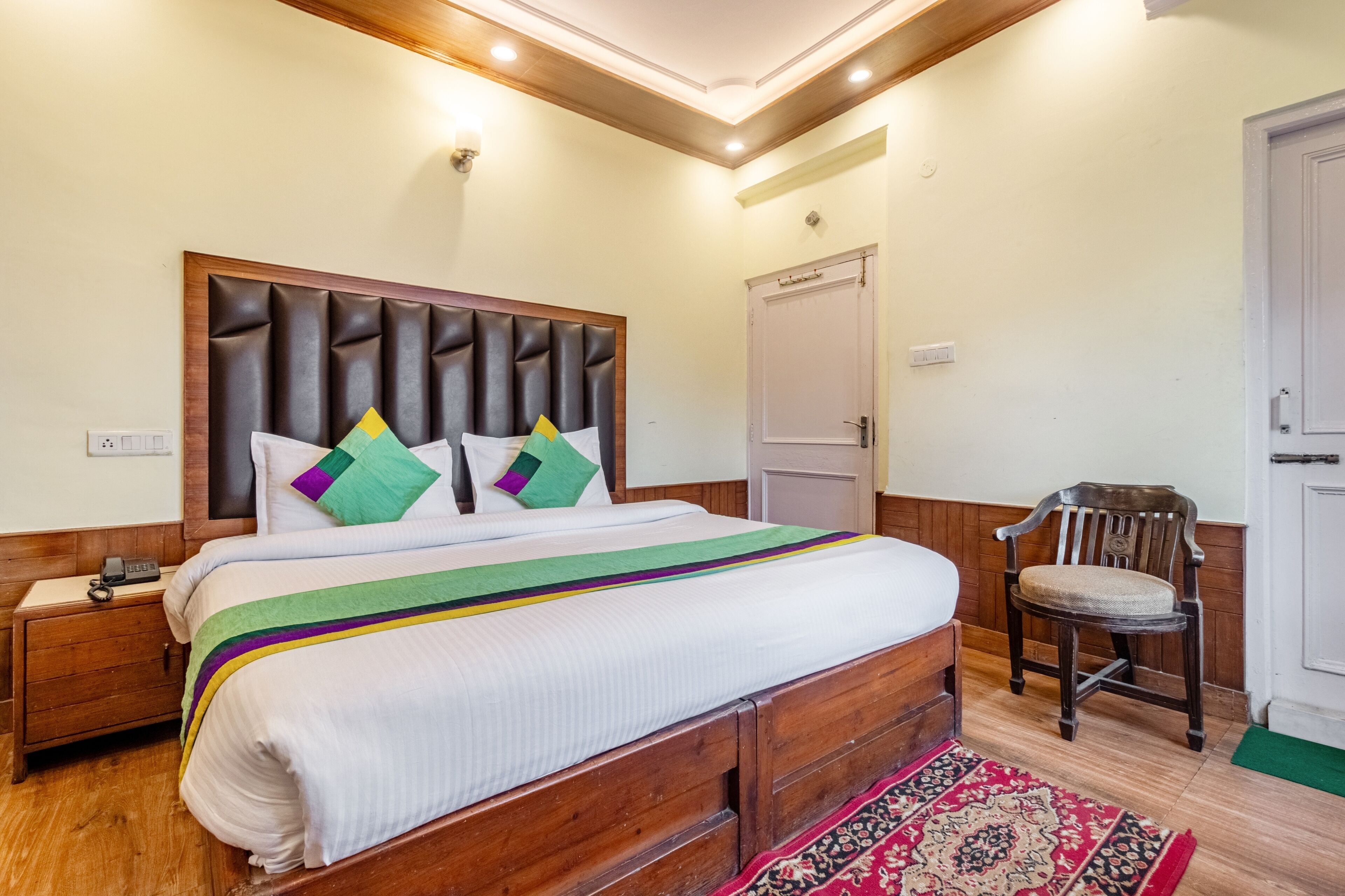A neatly made bed with a wooden headboard, a bedside table with a phone, a chair, and a door in a well-lit room.