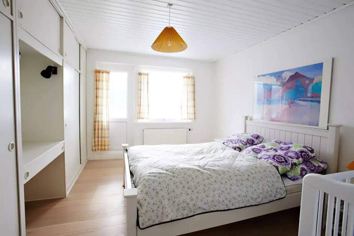 A bedroom with a bed, a wardrobe, a window with curtains, a radiator, and a painting on the wall.