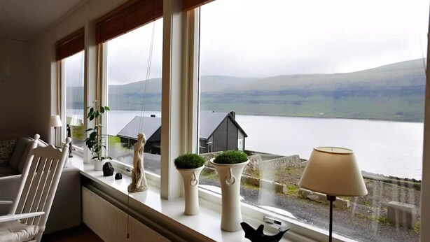 A room with a view of a lake and mountains, a white chair, a lamp, and decorative items on a windowsill. - Liljulon - Views - Location (Ljosa)