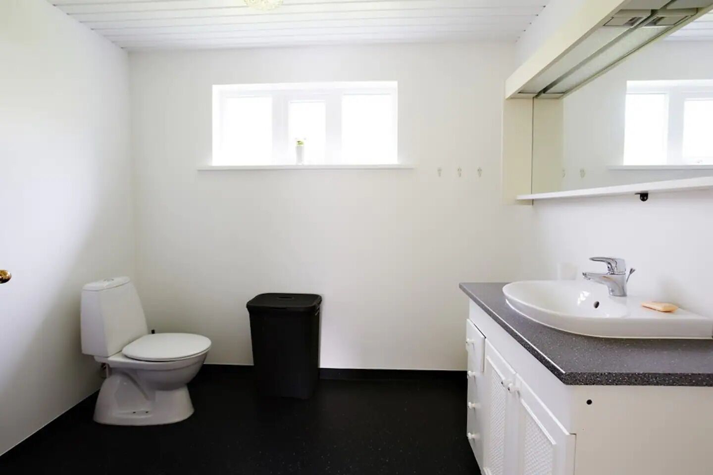 A small, clean bathroom with a toilet, a trash bin, a sink, and a window.