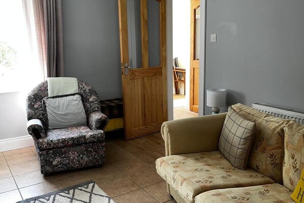 A living room with a patterned armchair, a sofa with floral cushions, a wooden door, and a lamp. - 3br-sea View-fireplace-garden-free Parking (Falcarragh)