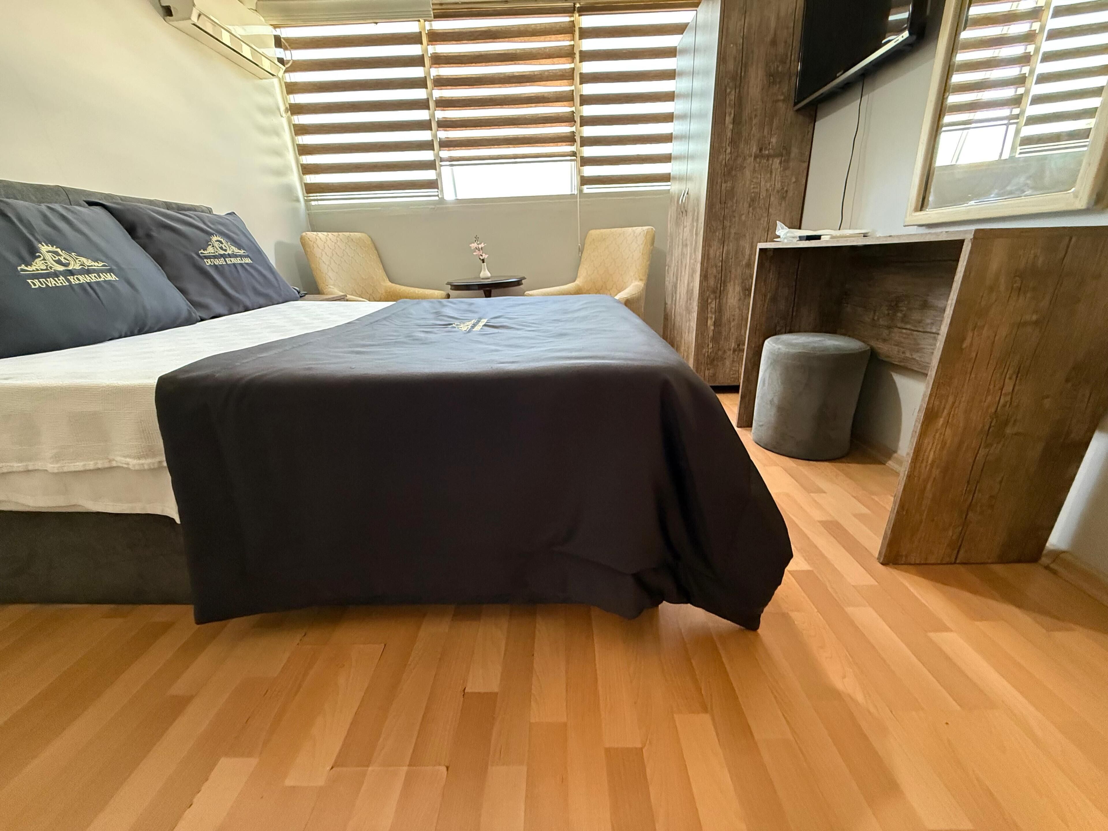 A bedroom with a bed, a small table, a chair, a wooden cabinet, and a window with blinds.