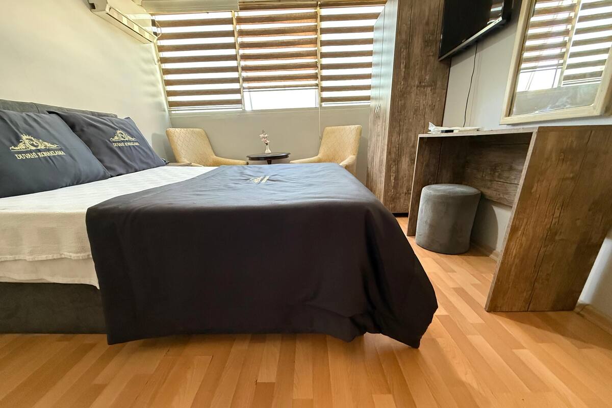 A bedroom with a bed, a small table, a chair, a wooden cabinet, and a window with blinds.