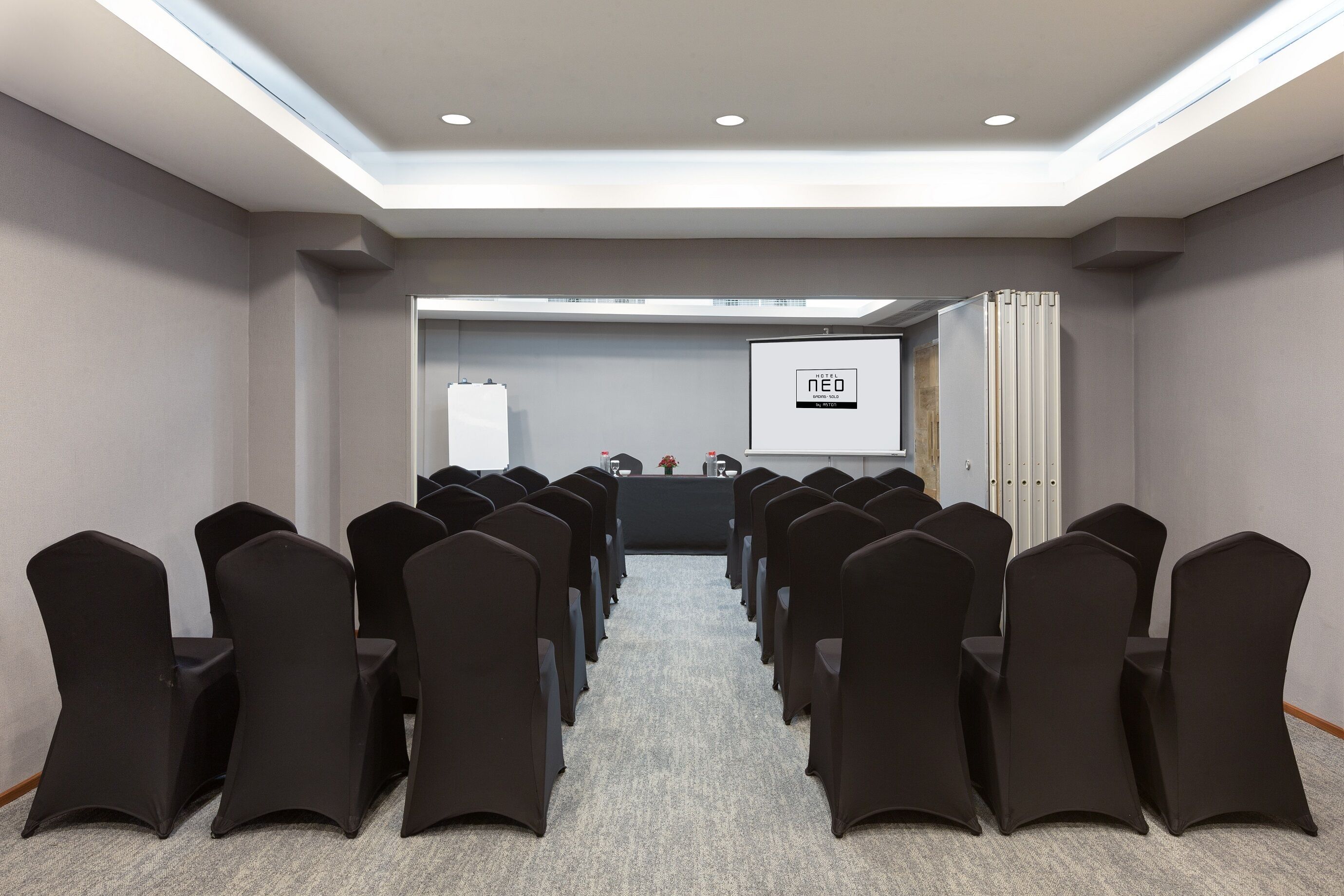 Un salón de conferencias con filas de sillas negras, una pantalla de proyector y una pizarra.