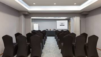 Un salón de conferencias con filas de sillas negras, una pantalla de proyector y una pizarra.