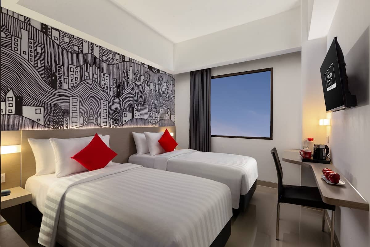 A hotel room with two beds, a desk, a chair, a TV, and a cityscape mural on the wall.
