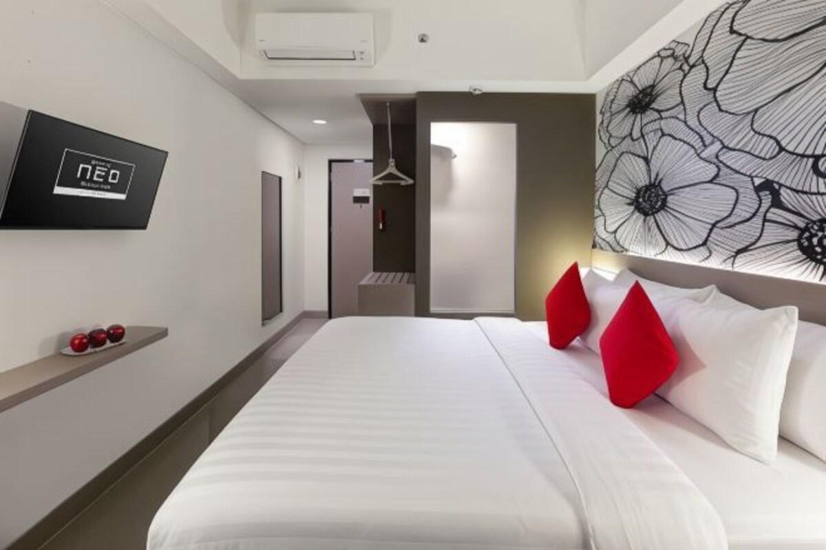 A modern hotel room with a large bed, red pillows, a wall-mounted TV, and a floral artwork.