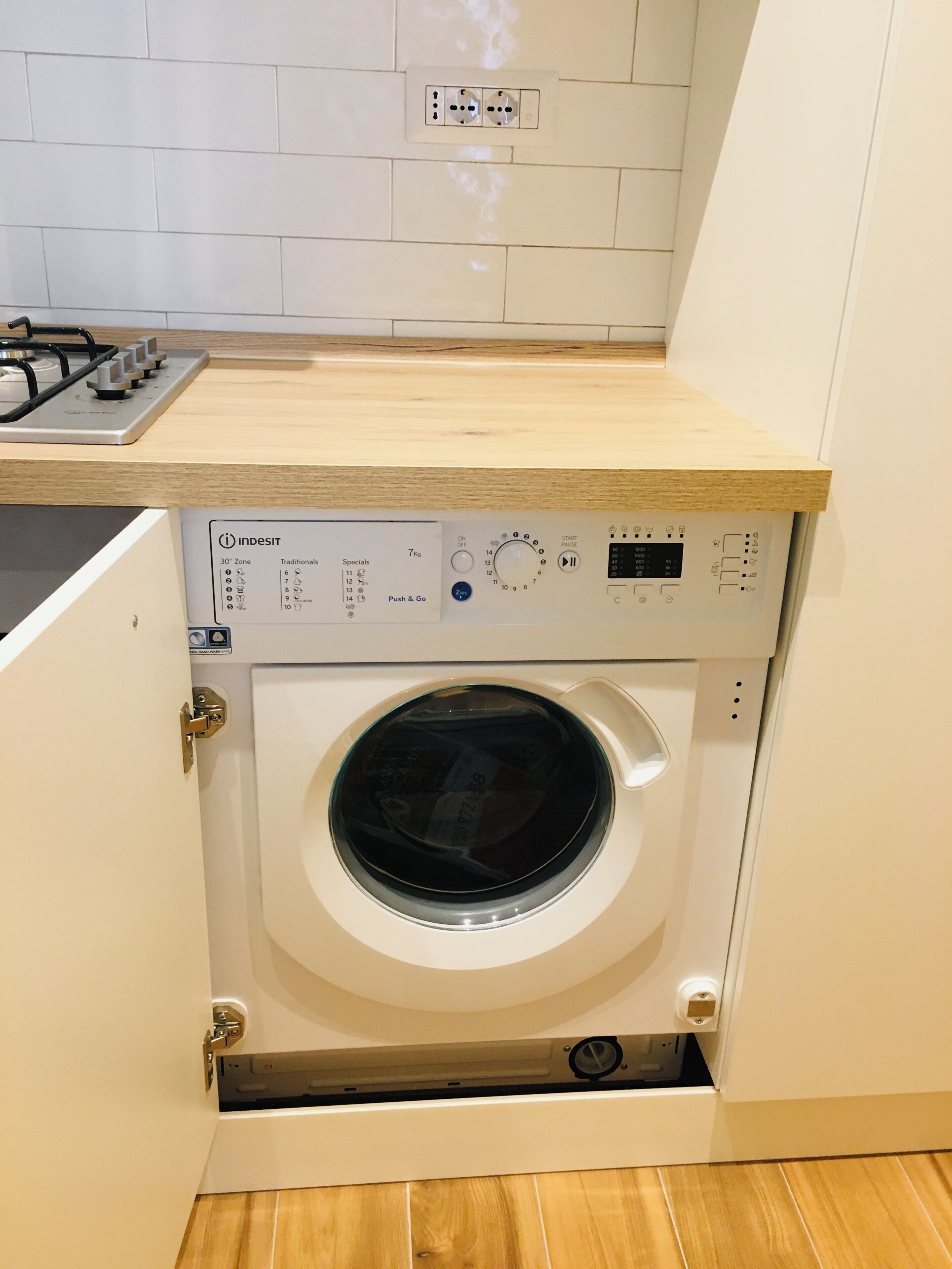 Eine kompakte Waschmaschine in einem kleinen Schrank mit Bedienfeld, einem Steckdosenanschluss darüber und einer Holz Arbeitsplatte.