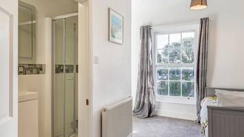 A bathroom with a shower, a radiator, a window with curtains, and a bed with a bedside table.