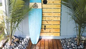 Ein Surfbrett, das an einer Holzwand angelehnt ist, mit einem dreieckigen Logo, umrahmt von zwei Palmen, und einem Holzboden davor.