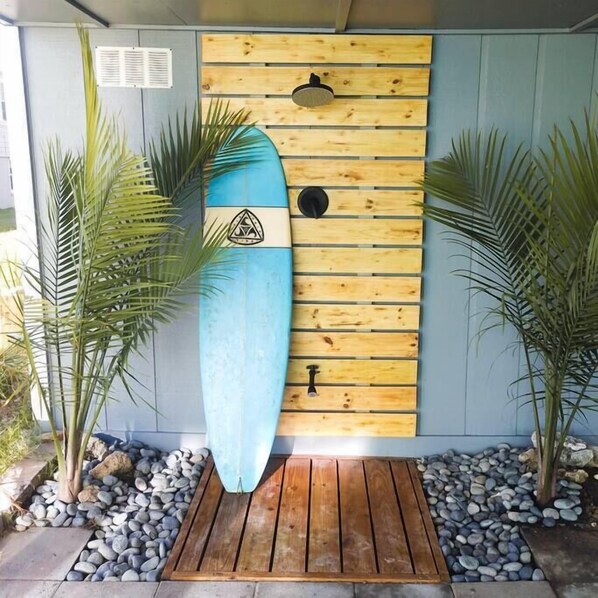 Una tabla de surf apoyada contra una pared de madera con un logotipo triangular, flanqueada por dos palmeras y una cubierta de madera al frente.