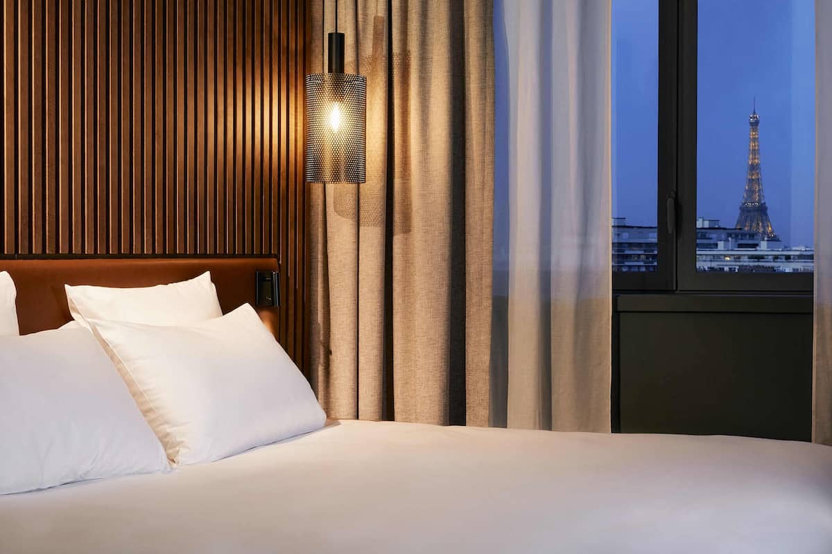 A hotel room with a bed, a nightstand, a lamp, and a view of the Eiffel Tower.